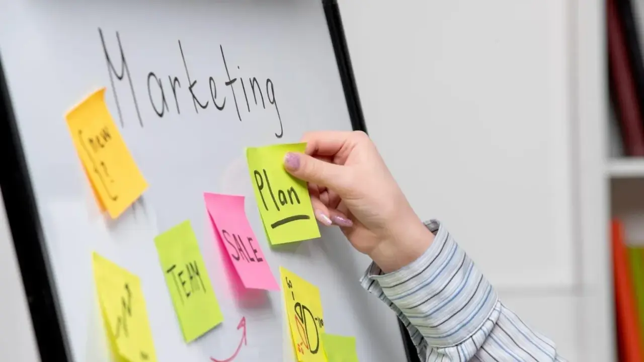 Ręka dodaje karteczkę z napisem "Plan" do tablicy z hasłem "Marketing". To przykład, jak crowdsourcing co to może pomóc w tworzeniu strategii.