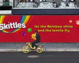 Skittles reklama: mężczyzna na rowerze pod tęczowym logo, latające cukierki. "Niech tęcza świeci, a soczewki latają".