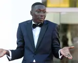 Khaby Lame, najpopularniejsza tiktokerka, w eleganckim smokingu z muszką, z gestem otwartych dłoni.