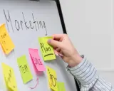 Ręka dodaje karteczkę z napisem "Plan" do tablicy z hasłem "Marketing". To przykład, jak crowdsourcing co to może pomóc w tworzeniu strategii.