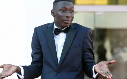 Khaby Lame, najpopularniejsza tiktokerka, w eleganckim smokingu z muszką, z gestem otwartych dłoni.