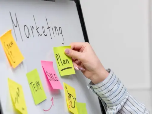 Ręka dodaje karteczkę z napisem "Plan" do tablicy z hasłem "Marketing". To przykład, jak crowdsourcing co to może pomóc w tworzeniu strategii.