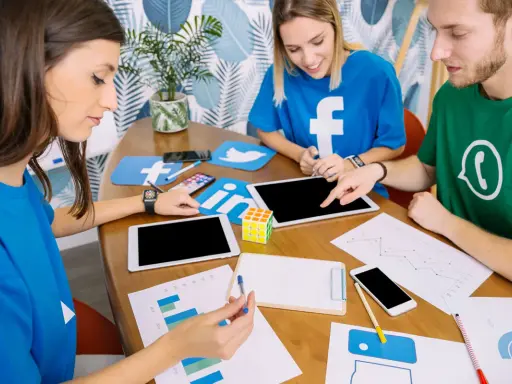 Trzy osoby przy stole omawiają, jak utworzyć grupę na Facebooku, otoczone logotypami mediów społecznościowych.