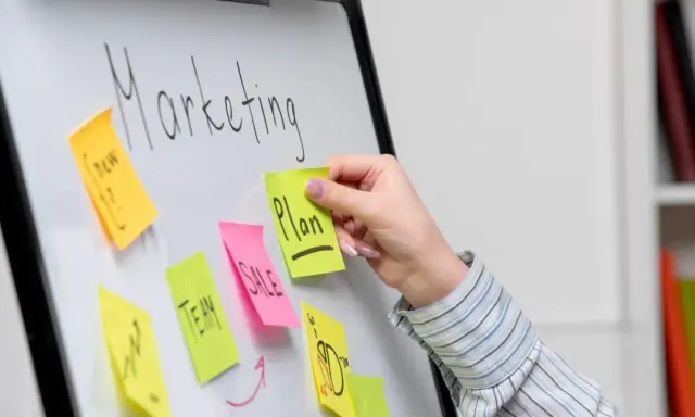Ręka dodaje karteczkę z napisem "Plan" do tablicy z hasłem "Marketing". To przykład, jak crowdsourcing co to może pomóc w tworzeniu strategii.