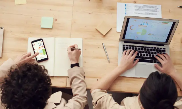 Dwie osoby analizują dane na laptopie i telefonie. Jedna pisze notatki, druga pracuje przy klawiaturze. To przykład pracy zespołowej, gdzie amp co to jest kluczowe dla sukcesu.